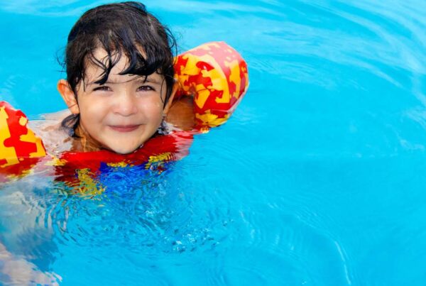 Niño pequeño con chaleco salvavidas siendo supervisado por un adulto en una piscina segura con valla perimetral.