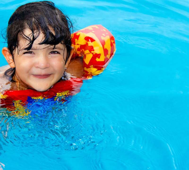 Niño pequeño con chaleco salvavidas siendo supervisado por un adulto en una piscina segura con valla perimetral.