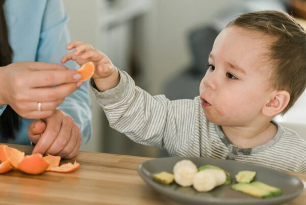 Niño pequeño comiendo fruta en trozos seguros mientras un adulto lo supervisa atentamente en la mesa.