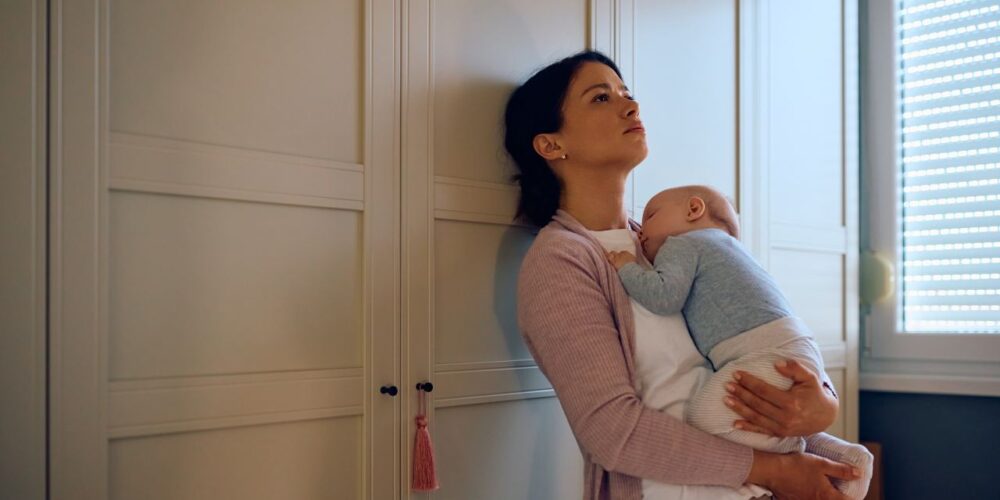 Madre con expresión de cansancio sostiene a su bebé mientras mira por la ventana, representando la depresión posparto prolongada.