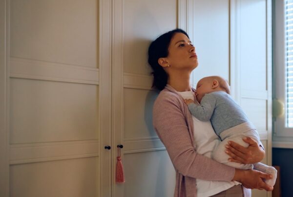 Madre con expresión de cansancio sostiene a su bebé mientras mira por la ventana, representando la depresión posparto prolongada.