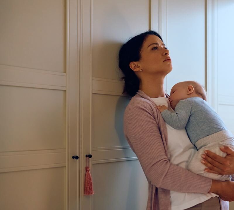 Madre con expresión de cansancio sostiene a su bebé mientras mira por la ventana, representando la depresión posparto prolongada.