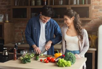 Hombre y mujer de mediana edad sonriendo mientras preparan una ensalada con vegetales frescos, representando una dieta saludable para la longevidad.