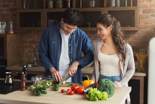 Hombre y mujer de mediana edad sonriendo mientras preparan una ensalada con vegetales frescos, representando una dieta saludable para la longevidad.