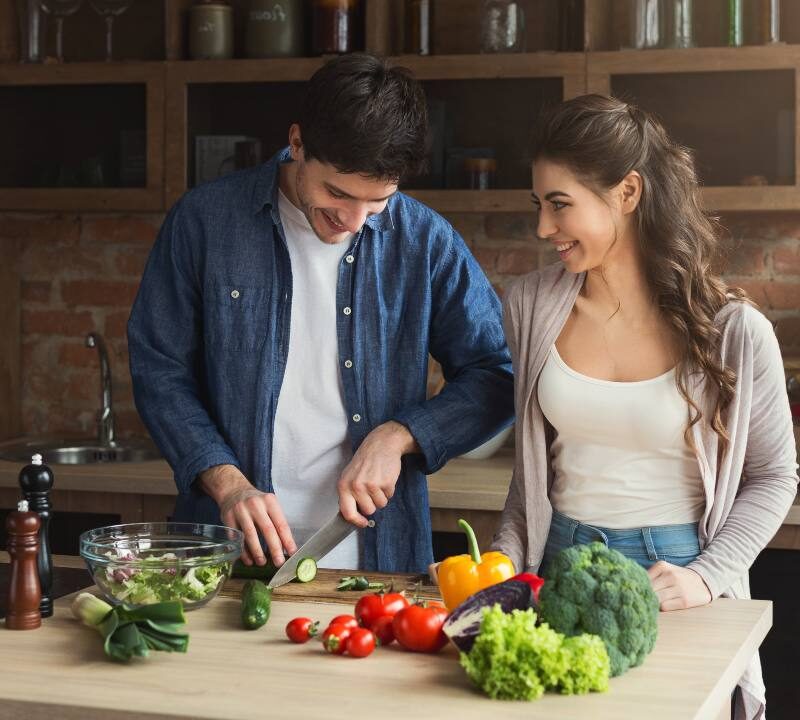 Hombre y mujer de mediana edad sonriendo mientras preparan una ensalada con vegetales frescos, representando una dieta saludable para la longevidad.