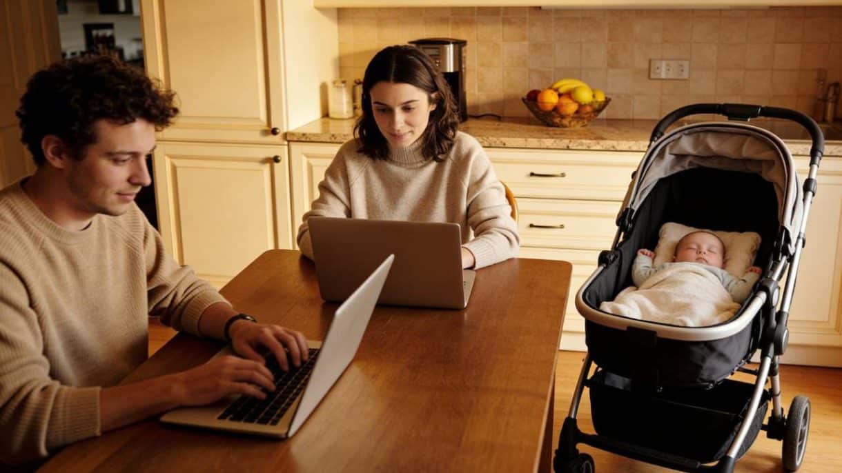 Una pareja joven trabajando con portátiles en el salón de su casa mientras un bebé descansa cerca, ilustrando la conexión entre teletrabajo y fertilidad.