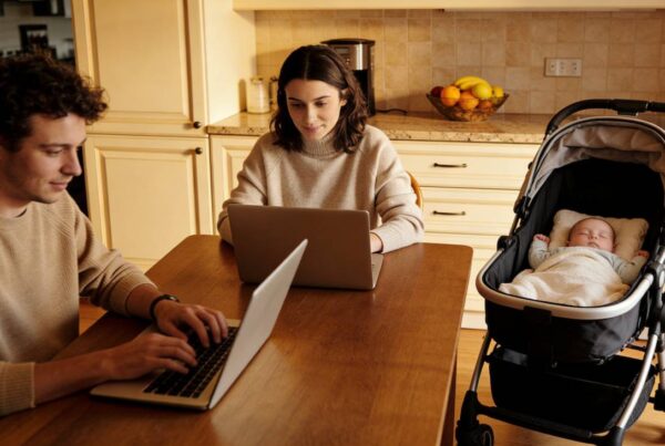 Una pareja joven trabajando con portátiles en el salón de su casa mientras un bebé descansa cerca, ilustrando la conexión entre teletrabajo y fertilidad.